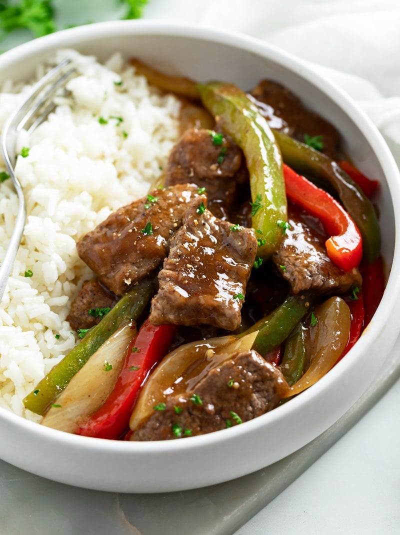 A white plate with Pepper Steak in a brown sauce next to white rice.