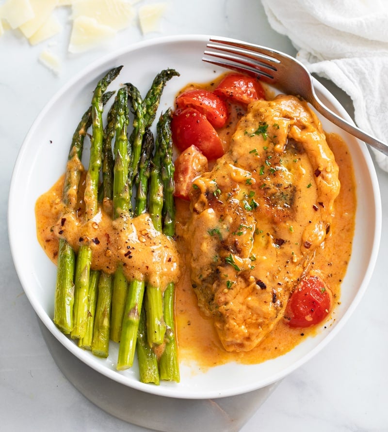 Creamy Tomato Chicken on a white plate next to asparagus with cherry tomatoes.