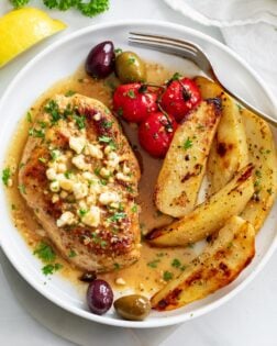 Greek Chicken on a white plate with Feta on top next to potatoes, tomatoes, and olives.