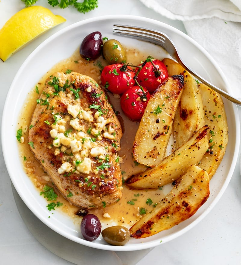 Greek Chicken on a white plate with Feta on top next to potatoes, tomatoes, and olives.