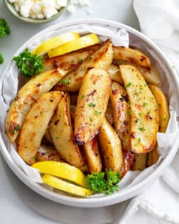 A white bowl of Greek Potatoes topped with parsley with lemon wedges on the side.