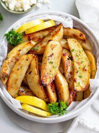 A white bowl of Greek Potatoes topped with parsley with lemon wedges on the side.