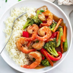 A white plate with shrimp stir fry in a brown sauce with rice and vegetables.