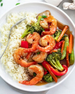 A white plate with shrimp stir fry in a brown sauce with rice and vegetables.