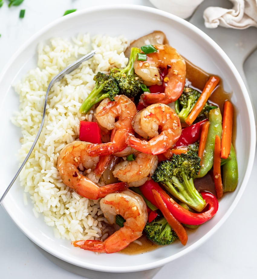 A white plate with shrimp stir fry in a brown sauce with rice and vegetables.