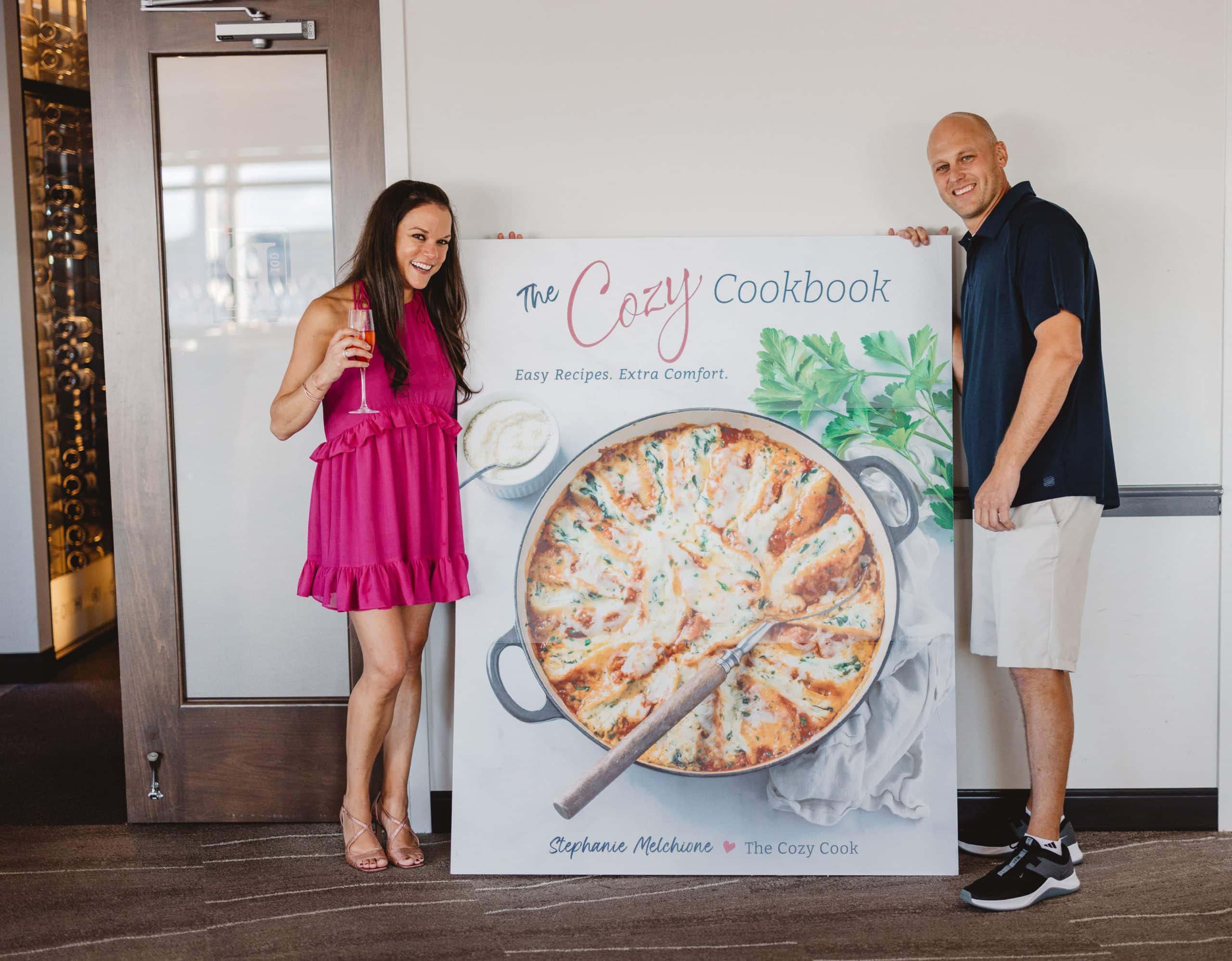 Two people standing next to a giant cover of The Cozy Cookbook.