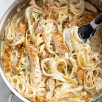 Chicken Alfredo Pasta in a stainless steel skillet with kitchen tongs, fettuccine, chicken strips, and a cream sauce.