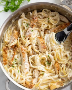 A skillet filled with Chicken Alfredo Pasta with fresh parsley and kitchen tongs in the skillet.