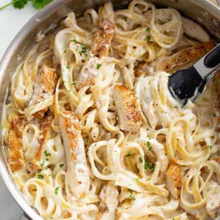 A skillet filled with Chicken Alfredo Pasta with fresh parsley and kitchen tongs in the skillet.