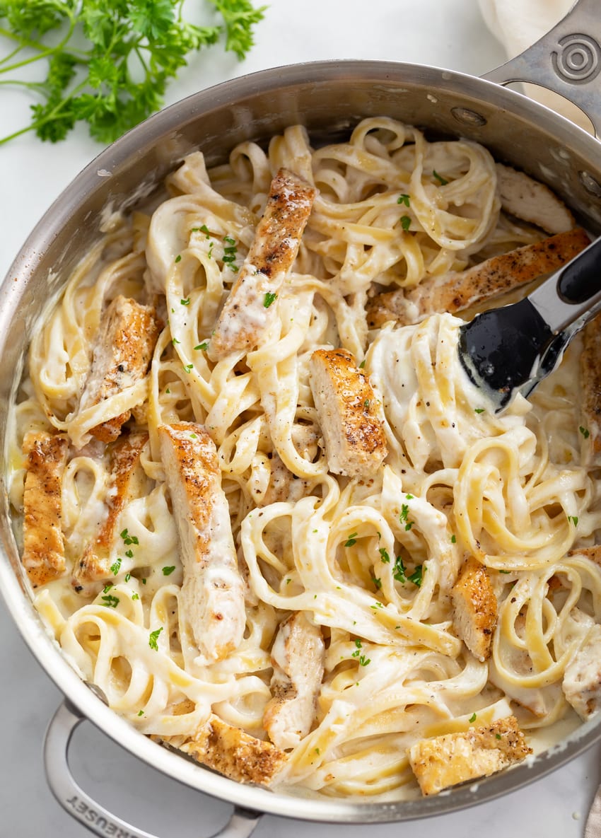 A skillet filled with Chicken Alfredo Pasta with fresh parsley and kitchen tongs in the skillet.