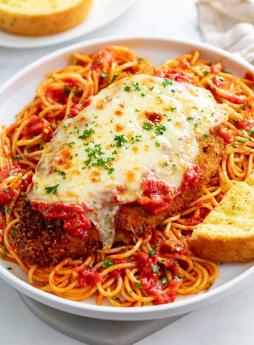 Chicken Parmesan on top of spaghetti with garlic bread on the side.