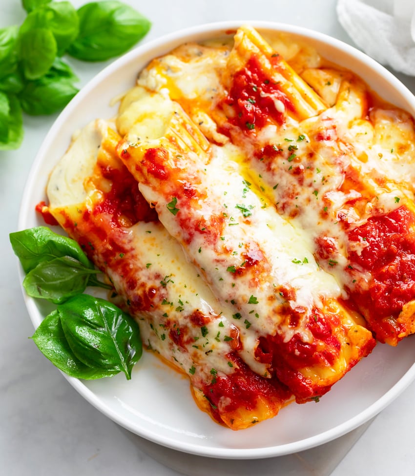 A white plate with manicotti on top with basil on the side.