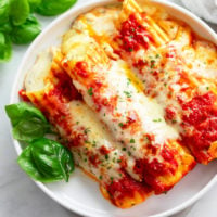 A white plate with Manicotti topped with sauce and cheese with basil on the side.
