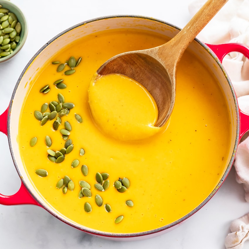 A red soup bowl filled with Butternut Squash Soup with pumpkin seeds on top and a spoon on the side.
