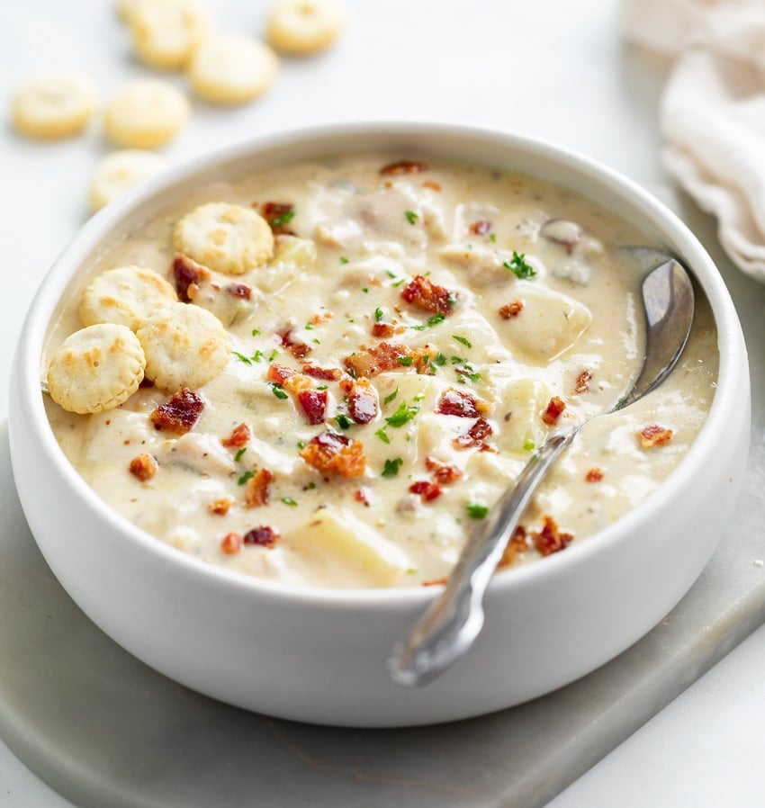A white bowl filled with New England Clam Chowder with bacon and oyster crackers on top.