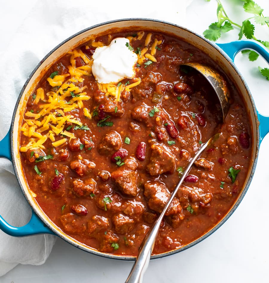 A Dutch oven filled with Chili Con Carne with shredded cheese, sour cream, and a ladle.