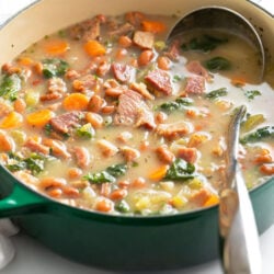 A green Dutch oven with Ham Soup with beans and vegetables with a ladle on the side.