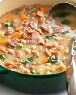 A green Dutch oven with Ham Soup with beans and vegetables with a ladle on the side.