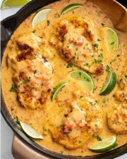 A skillet of Cajun Chicken in a sauce with cilantro and lime wedges.