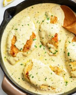 A skillet filled with Herb Chicken in a cream sauce with fresh parsley and a wooden spoon on the side.