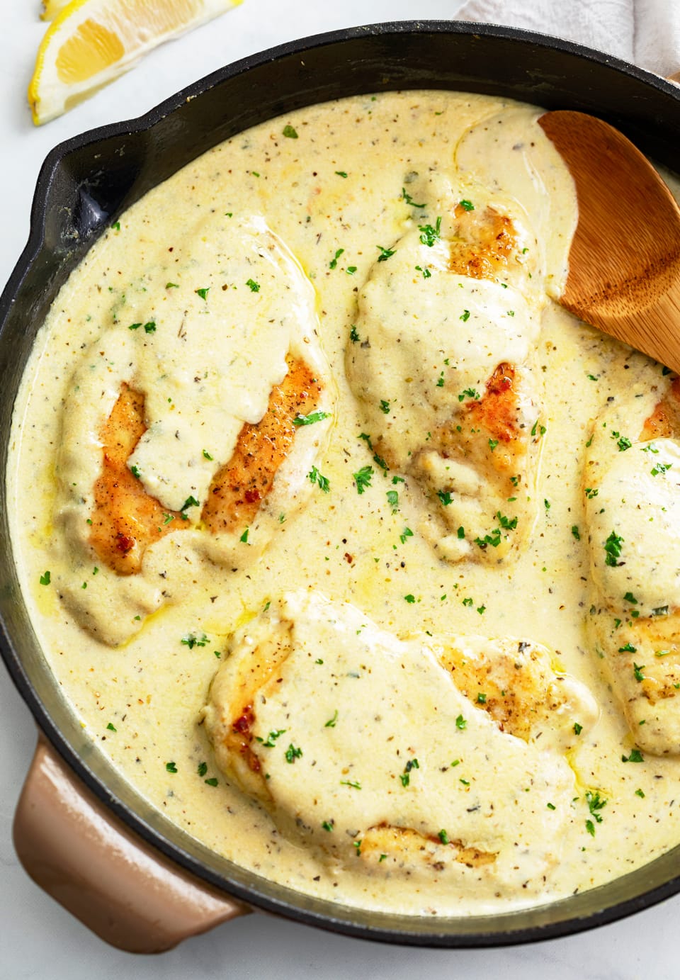 A skillet filled with Herb Chicken in a cream sauce with fresh parsley and a wooden spoon on the side.