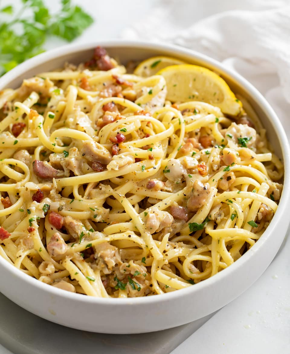 Linguine with Clams in a white bowl with pancetta, parsley, and lemon wedges.
