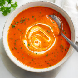 A bowl of Roasted Red Pepper Soup with cream swirled on top and a spoon on the side.