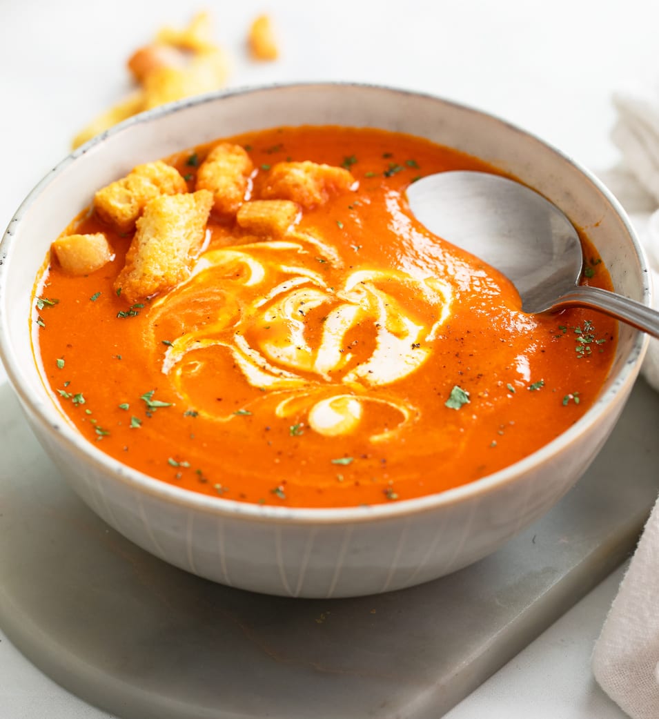 A white bowl filled with Roasted Red Pepper Soup with croutons on top and a spoon on the side.