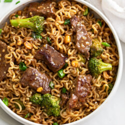 A white bowl with Ramen noodles and beef and broccoli in a brown sauce.