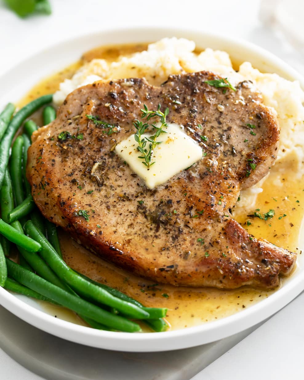 A Pan Fried Pork Chop on a plate with pan juice, mashed potatoes and green beans.