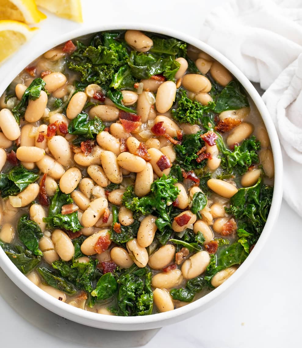 A white bowl with white beans and greens topped with chopped bacon.