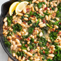 Greens and Beans in a skillet with lemon wedges on the side and bacon on top.