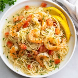 A white plate of Shrimp Scampi with sliced lemons and diced tomatoes.