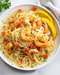 A white plate of Shrimp Scampi with sliced lemons and diced tomatoes.