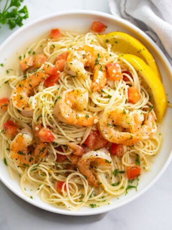 A white plate of Shrimp Scampi with sliced lemons and diced tomatoes.