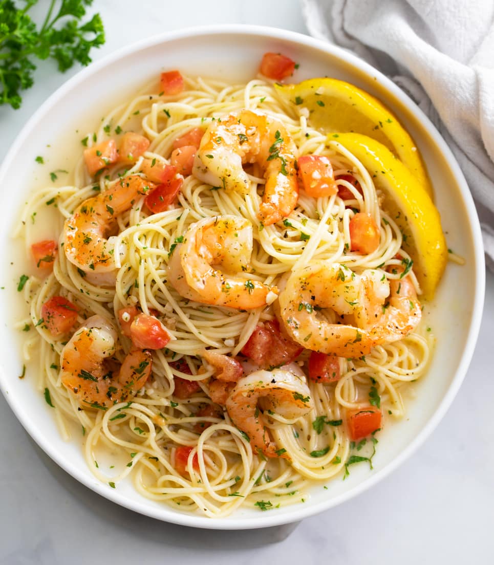 A white plate of Shrimp Scampi with sliced lemons and diced tomatoes.