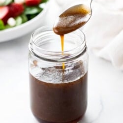 A spoon drizzling Balsamic Vinaigrette into a glass jar with a salad in the background.