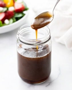 A spoon drizzling Balsamic Vinaigrette into a glass jar with a salad in the background.