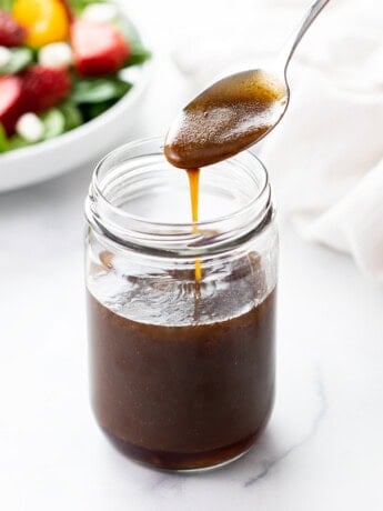 A spoon drizzling Balsamic Vinaigrette into a glass jar with a salad in the background.