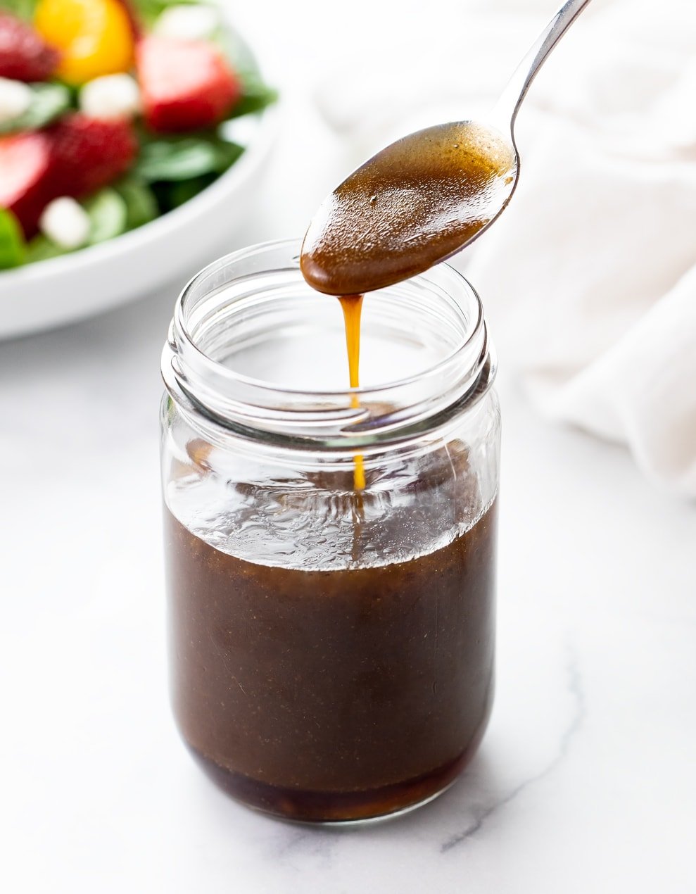 A spoon drizzling Balsamic Vinaigrette into a glass jar with a salad in the background.