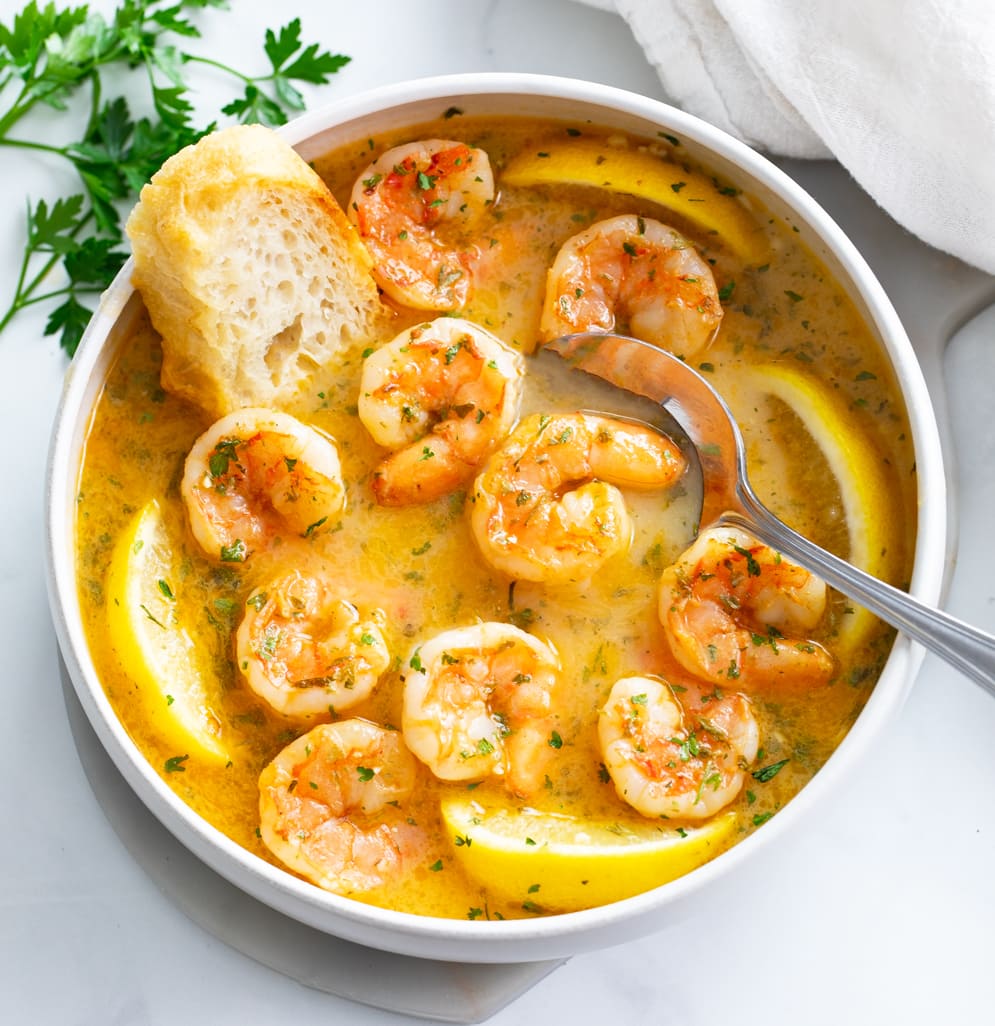 A white bowl with Garlic Butter Shrimp with a spoon and crusty bread on the side.