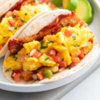 Breakfast Tacos filled with bacon, eggs, and hashbrown potatoes with avocado slices in the background.