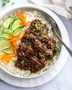 A white bowl of rice topped with Korean ground beef in sauce next to cucumbers and carrots.