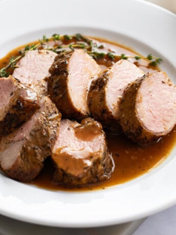 Slices of Roasted Pork Tenderloin on a white plate with pan sauce and thyme.