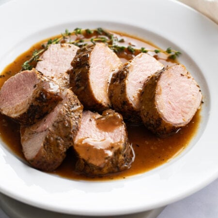 Slices of Roasted Pork Tenderloin on a white plate with pan sauce and thyme.