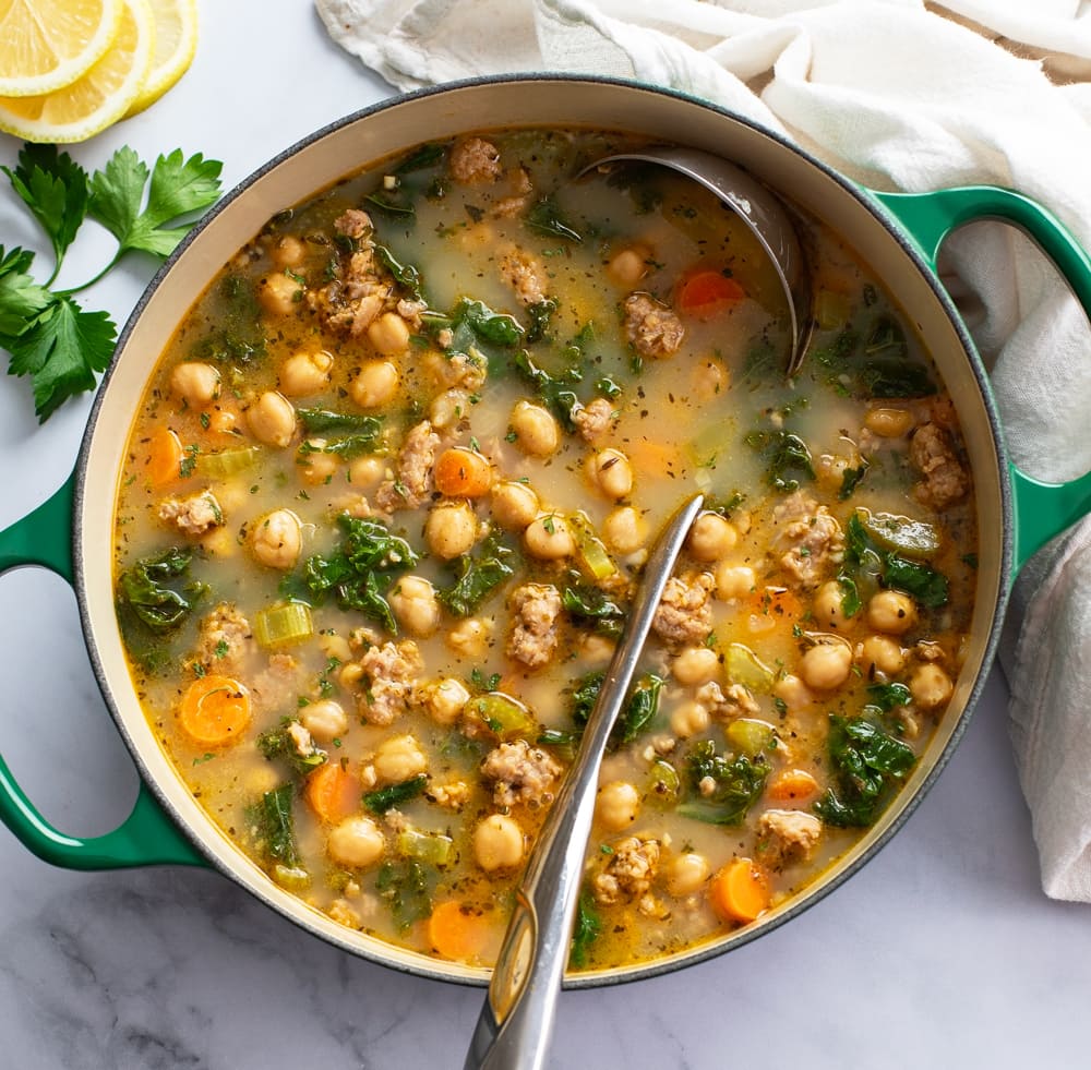 A pot of Chickpea Soup with a ladle in it and parsley and lemons on the side.