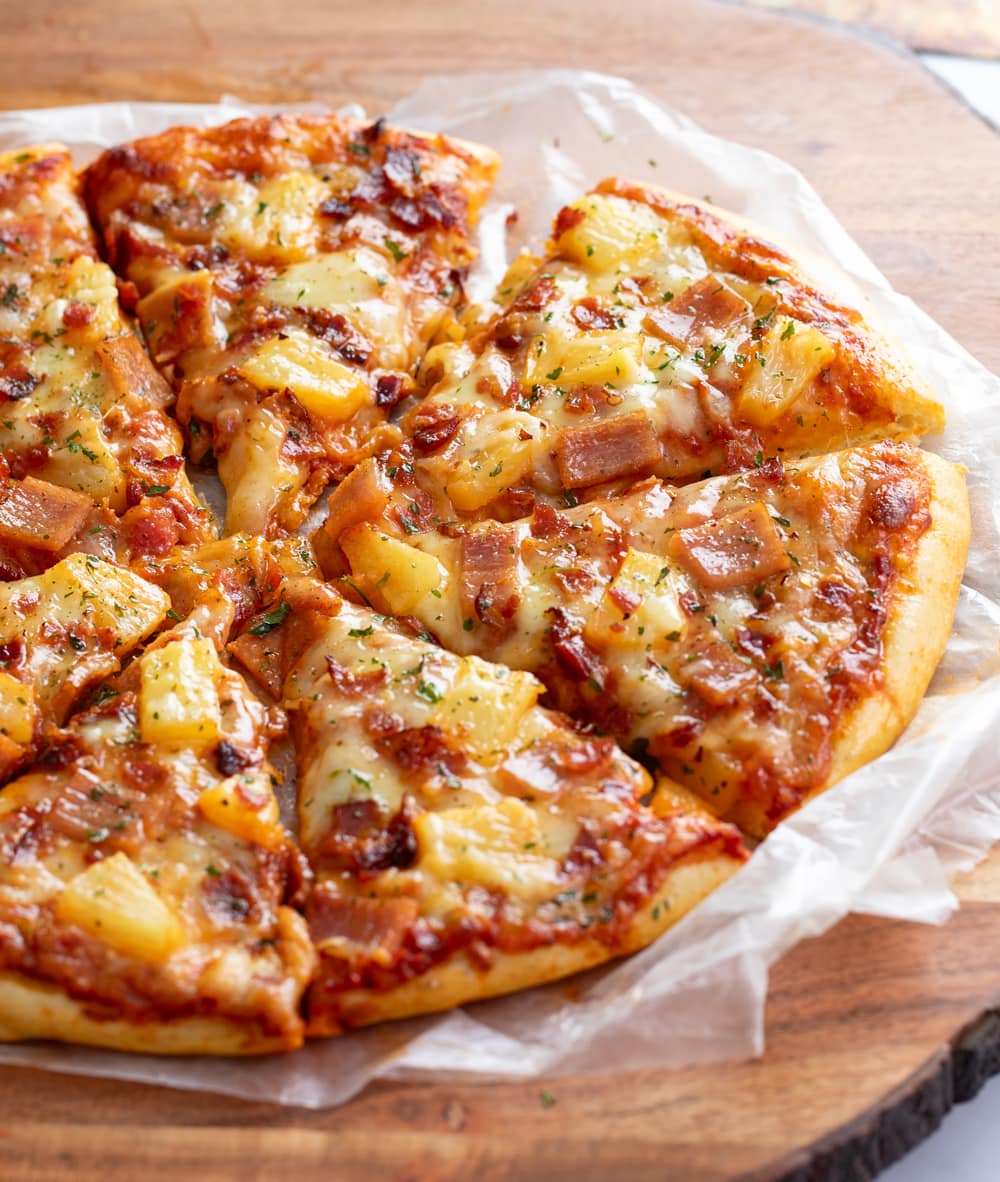 A Hawaiian Pizza on a wooden board cut into slices.