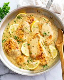 A skillet of Lemon Garlic Chicken with sauce, parsley, and lemons with a spoon on the side.