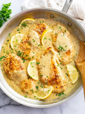 A skillet of Lemon Garlic Chicken with sauce, parsley, and lemons with a spoon on the side.