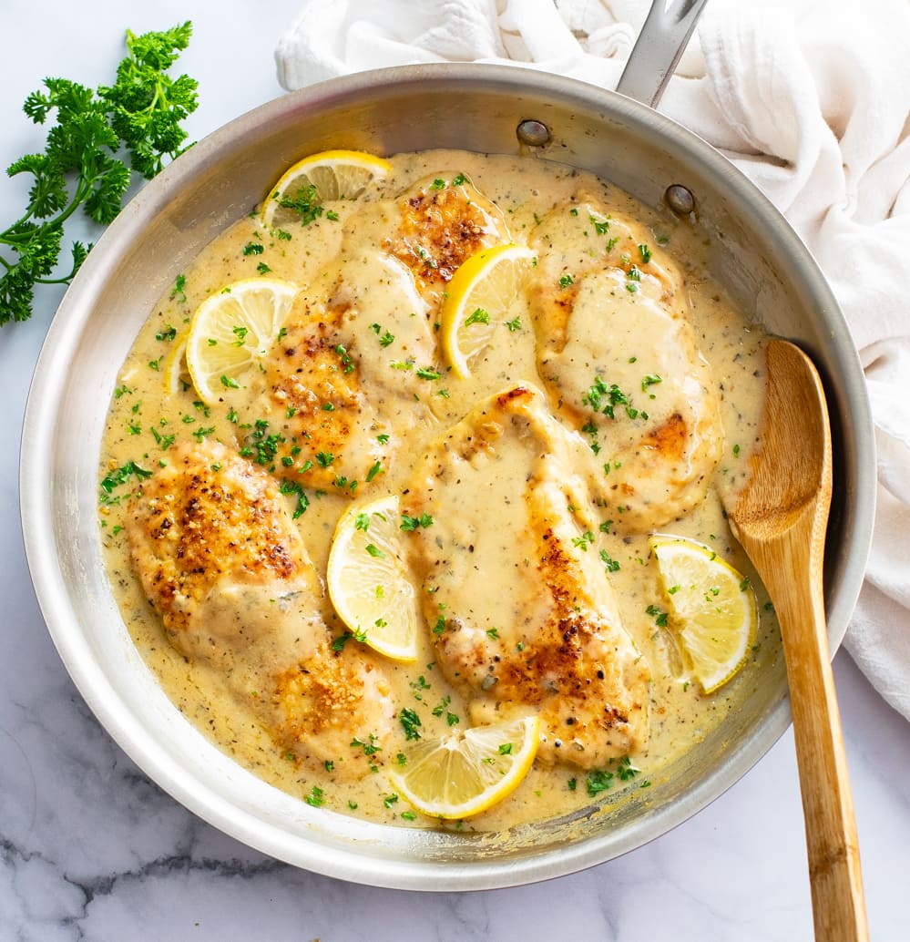 A skillet of Lemon Garlic Chicken with sauce, parsley, and lemons with a spoon on the side.